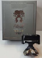 Photo showing the Pitbull Skull Shaver Gold Pro electric shaver in front of its grey branded box. The shaver is black with a gold accent and has a unique ergonomic shape.