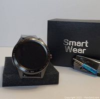 Photo of black metal smart watch on foam stand, Smart Wear branded box, and accessory in plastic packaging