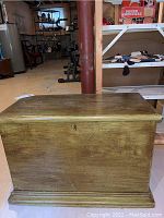 Photo of antique wooden blanket box from front angle showing wood grain and patina, lid closed, in basement setting.