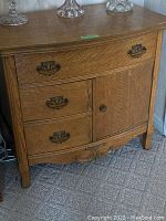 Front view of oak cabinet showing three drawers with ornate metal pulls and one door with knob, cabinet features carved bottom edge and curved top front edge.