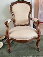 Front angled view of carved wood occasional chair showing carved wood frame, beige upholstery, and castor wheels.