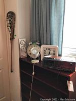 Lot displayed on wooden shelving showing wall clock, table clock, barometer, spotlamp, radio/cassette deck, and leaning lacrosse stick.