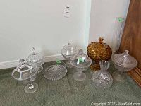 Seven pieces of glassware including six clear glass covered compote bowls, a clear glass vase, and one amber-colored covered bowl, displayed on green carpet.