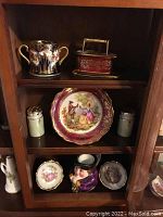 Side view showing porcelain cup with gold and floral design and decorative porcelain toggle with gold detail on top shelf, middle shelf plate with classical painted scene flanked by salt and pepper shakers, bottom shelf holding face-shaped mug and two small decorative plates
