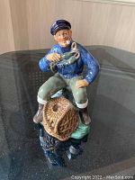 Front and side view of Royal Doulton The Lobster Man figurine showing fisherman holding lobster seated on rock and fishing net.