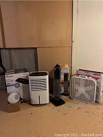 Wide view of all items: Lasko box fan, Bionaire tower heater/fan, Whirlpool dehumidifier, and light cover on floor against wall.