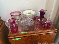 Seven cranberry and clear flash cranberry glass items arranged on a wooden surface, including pitcher, bowls, vase, pedestal bowl, and plate stand with decorative plate.