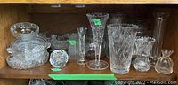Glass and crystal items displayed on a wooden shelf with various vases, bowls, candle holders, and other glassware.