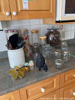 Entire lot displayed on kitchen counter showing multiple glass jars, metal tins, compote filled with pine cones, kitchen utensils in ceramic jar, and vintage irons.