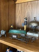 Shelf display showing bulldog figurine, man figurine, monkey bank, bell, and two candle snuffers.