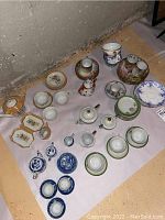Overall view of all miniature tea sets showing multiple tea cups, saucers, and teapots on a white surface near a textured wall.