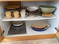Two shelves inside a wooden cabinet with stacked plates, mugs, bowls, and two ceramic baking dishes on lower shelf.