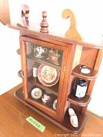 Front view of smaller wood and glass miniature collection shelf showing glass door with small displayed items inside and side open shelves with items.