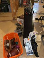 Shows weight bench with resistance bands attached, dumbbells, and orange basket with baseball gloves, footballs, and ice shoes on floor in basement.