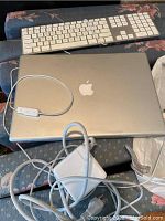 Top view of closed silver Apple MacBook laptop with attached charger and wired Apple keyboard in background