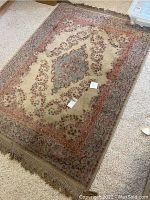 Overall view of one of the area rugs showing central medallion design with blue, pink, and cream colors and beige fringe edges, lying on carpeted floor.