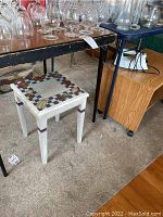Small white side table with mosaic tile top seen under a larger table with glassware on top.