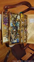 Wooden jewelry chest open displaying multiple compartments filled with assorted colorful costume jewelry including necklaces, pins, and bracelets.