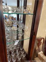 Overall display of crystal stemware set on three glass shelves inside a wooden glass cabinet showcasing various sizes and styles of glasses.