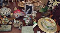 Photo showing assortment of figurines, Waterford crystal picture frame, porcelain dishware, napkin rings, and faux flowers on table