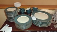Stack of dinner, salad, and bread plates in blue-green and cream with silver rim, cups, and saucers arranged on table.