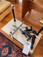 Fly tying stand base with metal rods and clamps on wooden floor with patterned rug corner visible.