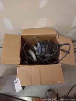 Open cardboard box containing a corded Skilsaw and two corded electric drills with tangled cords.