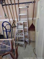 Two aluminum ladders leaning against a wall with garden tools and a refrigerator next to them.