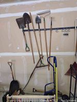 Photo showing multiple shovels, garden rake, sledgehammer, and a garden hoe hung on a wall rack in a garage.