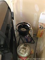 Photo showing outdoor propane burner stove, propane gas canister with red label, bag of fuel, and propane-labeled cardboard box in a garage corner