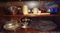 Nambe silverplate bowl on shelf with electric candles and blue and red plastic flatware storage containers above. Cloth bags and silverplate trays and candleholders below.