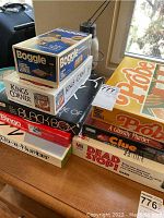 Stack of vintage board games including Boggle, King's Corner, Black Box, Bingo, and Do-a-Number seen on a wooden table near a window.