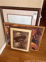 Four framed art pieces stacked against a wall: one limited edition signed pencil drawing of a person with a hat, three other framed artworks with various photographic or printed images