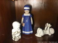 Three porcelain figurines: blue dress girl standing, seated child holding flowers, lying child with flowers on feet