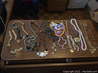 Wide view of various necklaces and pins laid out on a table including wooden bead necklaces, metal chains, and pin collection.