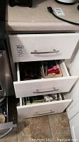 Three kitchen drawers partially open showing various kitchen utensils and supplies inside.