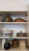Photo of a cabinet shelf containing seven pottery items including a greenish-brown pot, white ceramic dish with blue patterns, small brown jar, cream container, and dark mug with metal lid.
