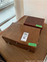 Photo of the two wooden jewelry boxes closed on a wooden table near a window, showing the smooth lid of the larger box and the carved lid of the smaller box.