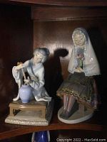 Photo showing both Lladro figurines side by side on a wooden shelf; the seated lady figurine to the right has a broken flower in her bouquet.
