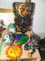 Carved wooden mask with multi-color paint and sun/moon face detail on the forehead surrounded by ceramic colorful birds, frog and sun face wall hanging.
