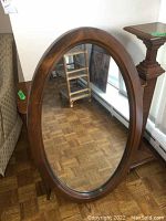 Full view of the oval mirror leaning against a chair and radiator, showing the wood grain frame and reflection of parquet flooring.