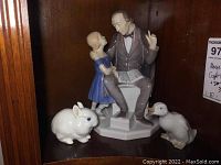 Overall view of the three porcelain figurines placed on a wooden shelf including the B&G Hans Christian Andersen and child figurine, Royal Copenhagen rabbit, and Lladro duck.