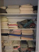 Closet shelf with multiple stacks of linens including folded towels, blankets, and linens in beige, cream, and pastel colors.