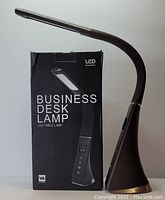 Photo shows a brown LED business desk lamp next to its original black box packaging labeled 'Business Desk Lamp' with an image of the lamp and text indicating LED table lamp and VA screen.