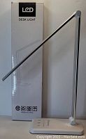 LED desk lamp standing upright next to its packaging box labeled 'LED Desk Light'. Lamp has a thin, adjustable arm and base with touchscreen controls.
