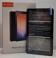 Tablet next to product box showing front and rear cameras with visible protective stickers and instruction text on the back.