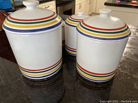 Four differently sized white ceramic kitchen canisters decorated with horizontal colorful stripes in red, yellow, green, and blue, with fitted lids.