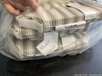 Photo showing a stack of four striped seat cushions in taupe/grey and off-white colors, packaged in a clear plastic bag, with visible velcro straps at the corners.
