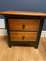 Front view of wooden bedside table showing two drawers with black knobs and black frame