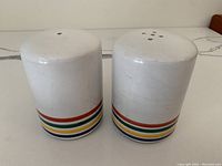Two white ceramic salt and pepper shakers side by side with colorful stripes at the base, showing tops with holes and crackle glaze on the surface.